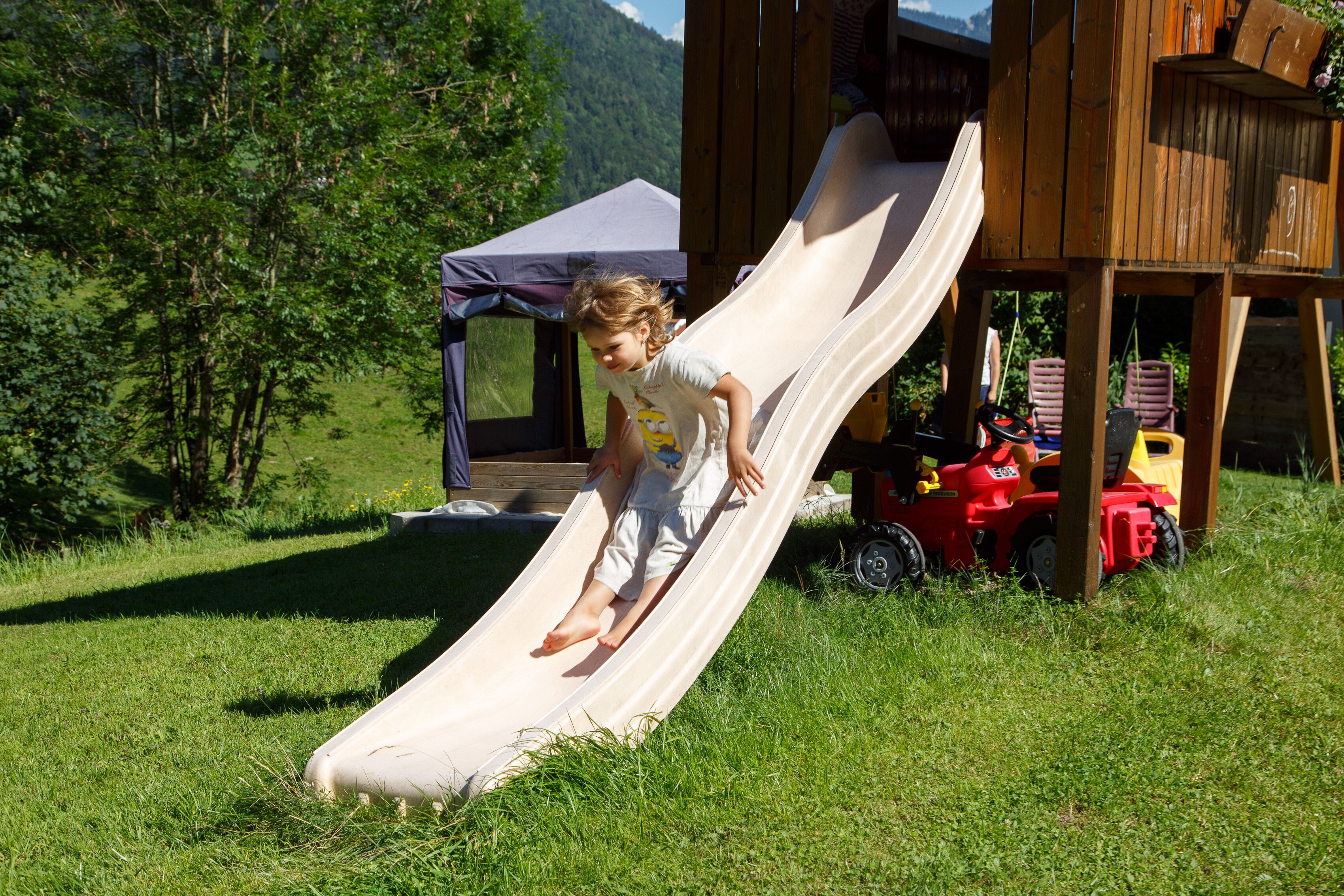 A child sliding down a slide in a grassy area with a wooden playhouse and a tent in the background.
