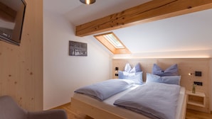Un dormitorio con techo de madera, un tragaluz, cama con ropa de cama azul, mesita de noche y televisor montado en la pared.