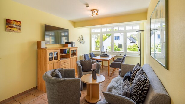 A living room with a sofa, chairs, a table, a TV, wooden cabinets, and a window with a view. - Apartment 013 / 1st floor with balcony/ Only bookable for 4 persons - Villa Meernixe - (Binz)