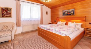 Une chambre confortable avec un plafond en bois, un grand lit avec une literie à motifs floraux, une table de chevet en bois, une chaise et un tableau accroché au mur.