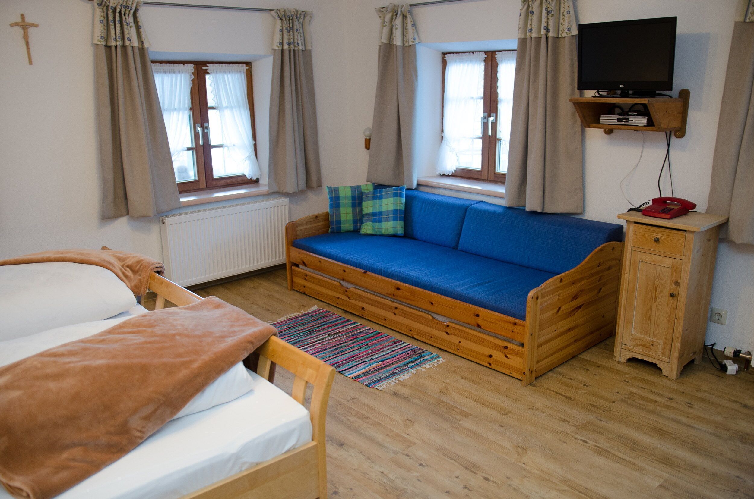 A room with a bed, a blue sofa, a wooden cabinet, a television on a shelf, and a window with curtains.