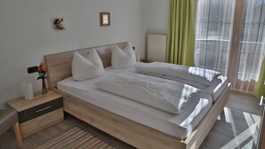 Een slaapkamer met een houten bed, witte beddengoed, een nachtkastje met een bloemenvaas, een ingelijst schilderij aan de muur, en een raam met groene gordijnen.