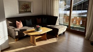 Een woonkamer met een houten salontafel, een bruine leren bank met kussens, een bloemschilderij in een lijst aan de muur en uitzicht op een besneeerd landschap door een glazen deur.