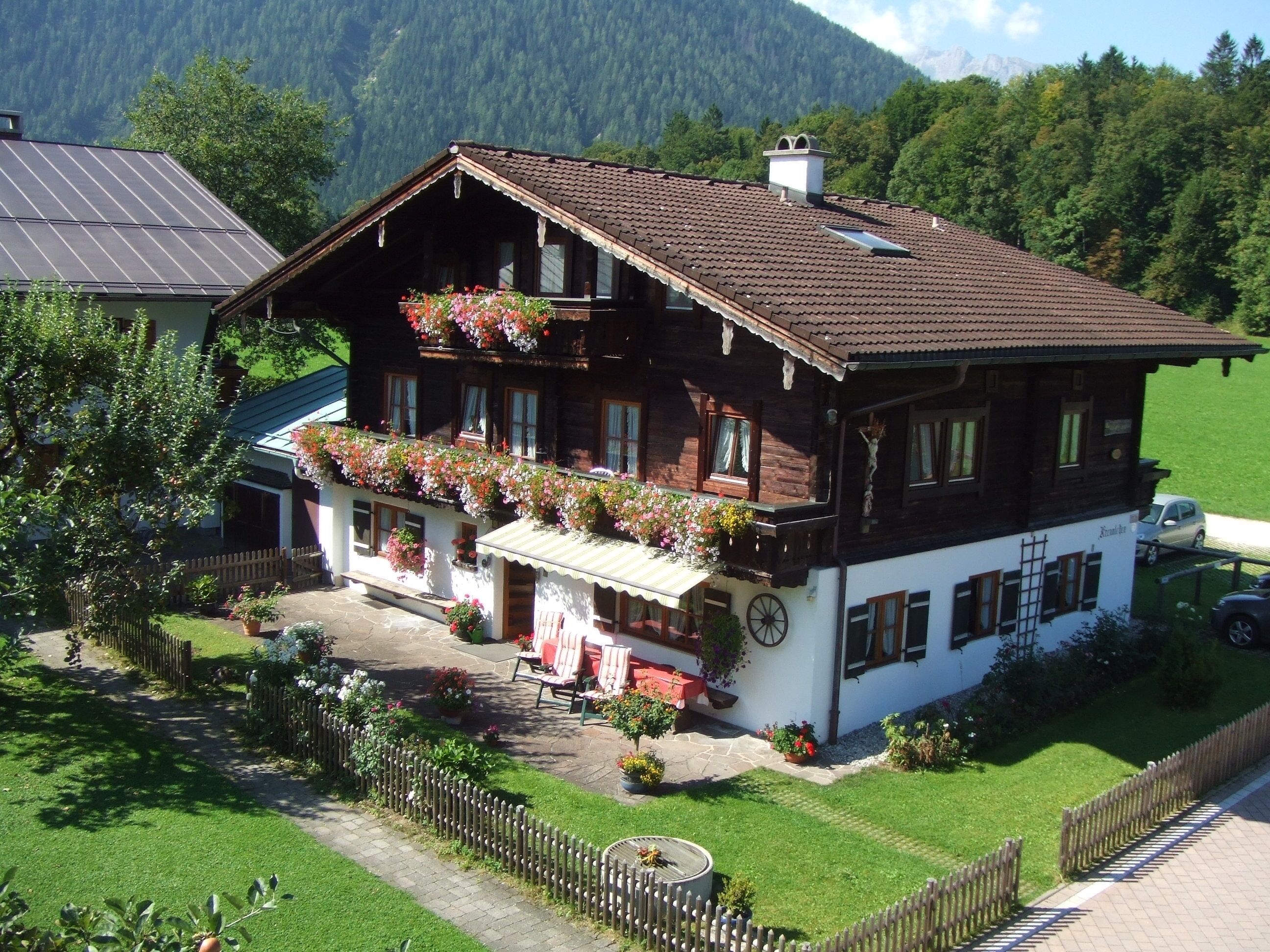 A traditional wooden chalet with a balcony adorned with flowers, a stone pathway leading to the entrance, and a view of the surrounding greenery and mountains.