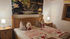 Un lit en bois avec une literie à motifs floraux, une table de chevet avec une lampe et un tableau représentant de l’eau et des fleurs accroché au mur.