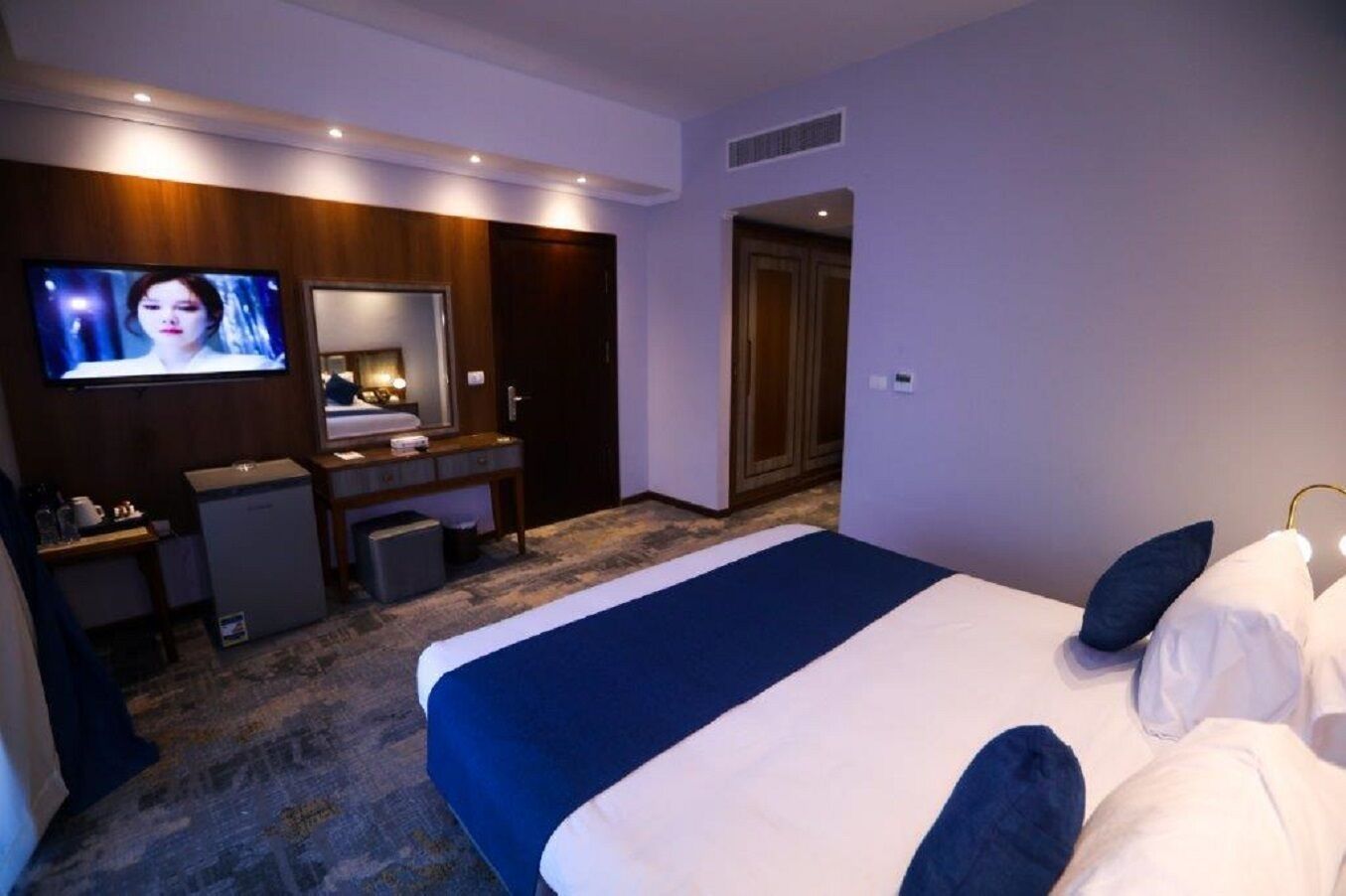 Une chambre d’hôtel avec un grand lit, une télévision, un bureau avec un ordinateur, un miroir et une armoire.