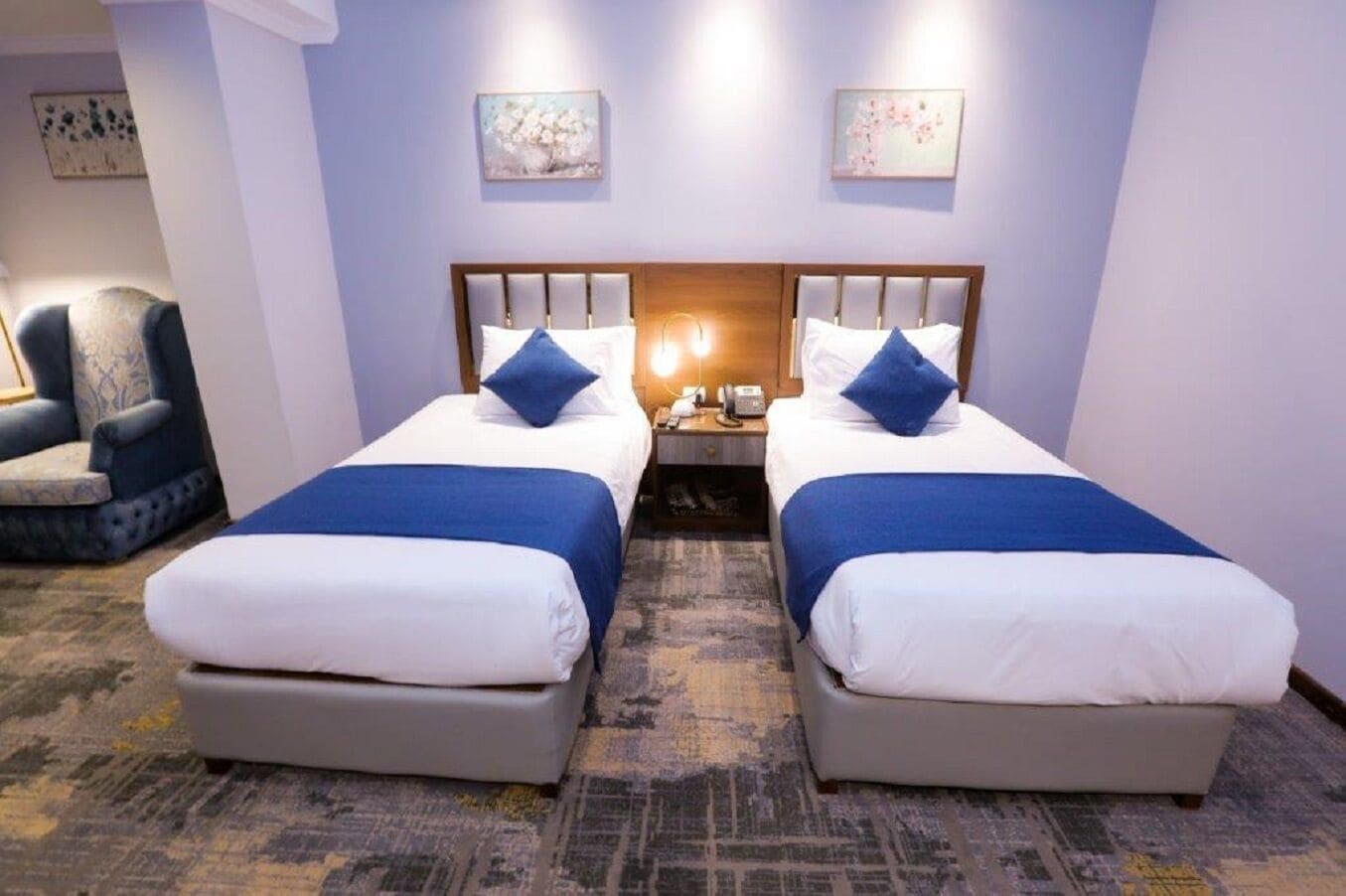 Habitación de hotel con dos camas, cada una con una almohada azul decorativa y una colcha blanca. Entre las camas, hay una mesita de noche con una lámpara y un teléfono. La habitación tiene una alfombra estampada y un sillón azul en la esquina.