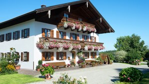 En traditionell chalet med en balkong som är prydd med blommor, en asfalterad uppfart och en trädgård med planterade blommor i krukor.