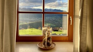 Ein Fenster mit freiem Blick auf eine gebirgige Landschaft, ein Holzrahmen und ein dekorativer Glasbehälter mit Kiefernzapfen auf einem Fensterbrett.