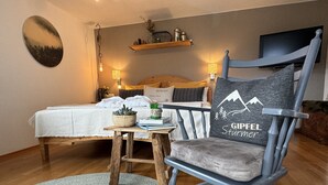 Ein gemütliches Zimmer mit einem Bett, einem Holztisch mit Pflanze, einem Stuhl mit Kissen mit Bergmotiv und einem Wand-Fernseher.