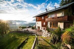 Un chalet de dos pisos con balcón, rodeado de abundante vegetación y montañas al fondo.