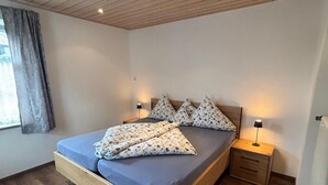 Une chambre à coucher avec un cadre de lit en bois, un matelas bleu, des coussins à motifs floraux, une table de chevet avec une lampe et une fenêtre avec des rideaux.