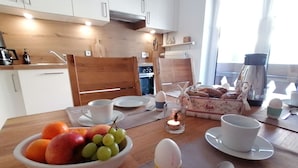 Une table de petit-déjeuner avec un bol de fruits, des œufs et du pain, placée sur une table en bois dans une cuisine aux armoires blanches et dotée d’une fenêtre.
