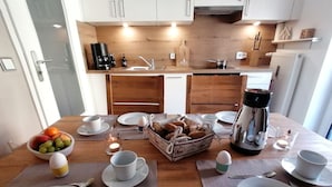 Un coin repas avec une table en bois mise pour le petit-déjeuner, comprenant des assiettes, des tasses et un panier de fruits.
