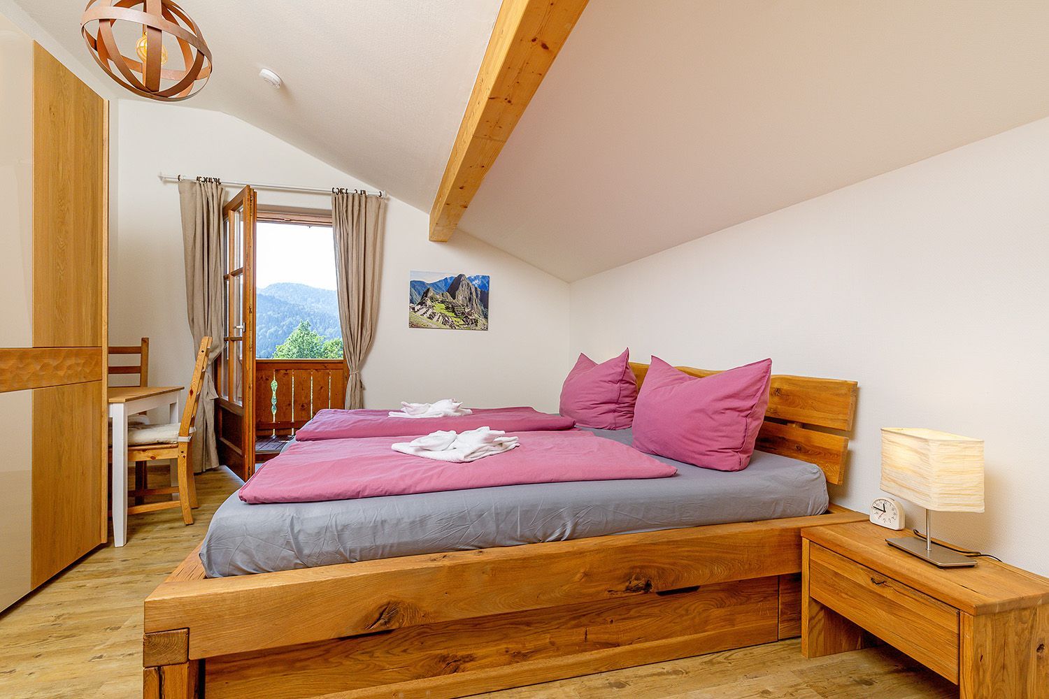 A bedroom with a wooden bed, a desk, a chair, a lamp, and a view of mountains through a window.
