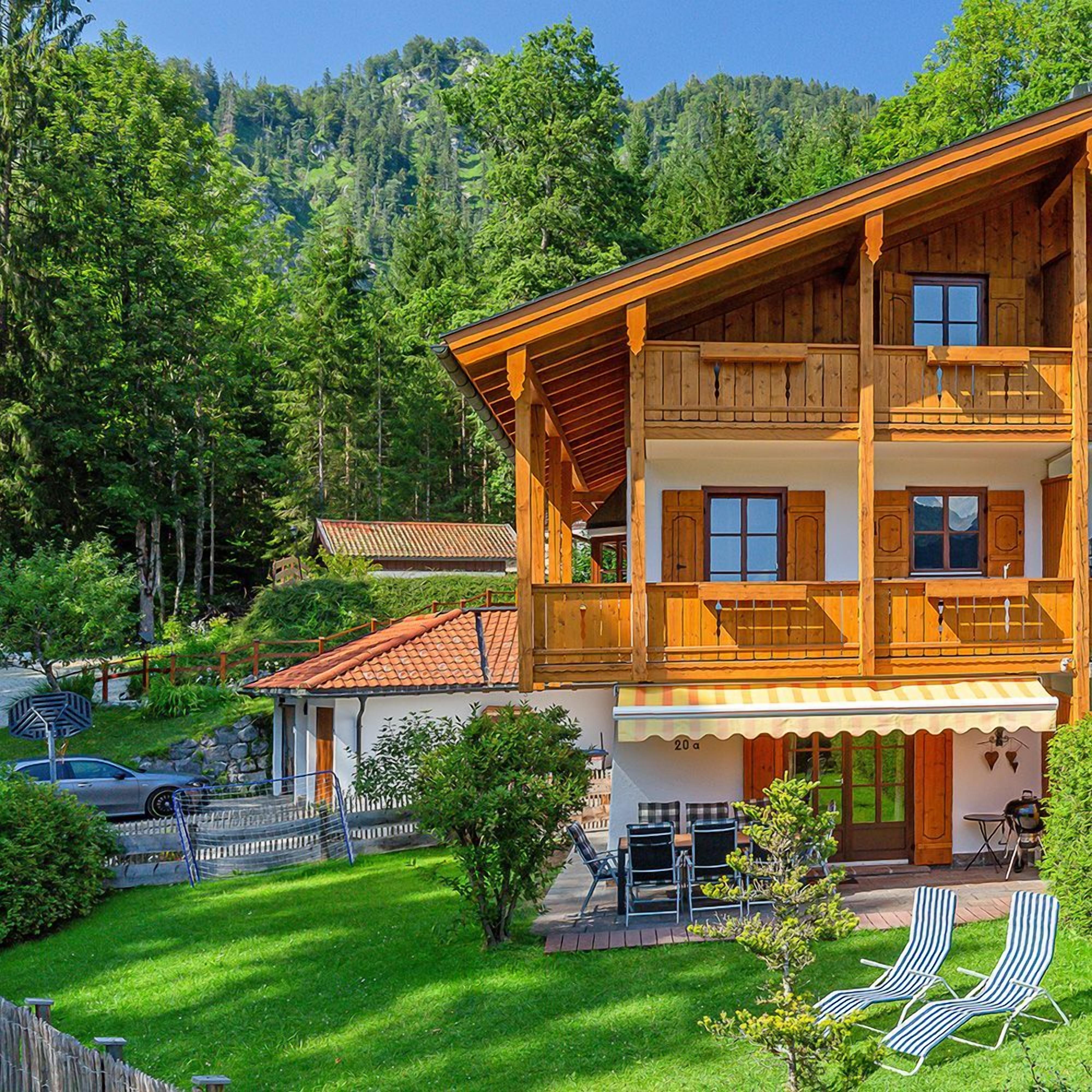 A two-story wooden chalet with a balcony, a covered patio area, and a view of a forested hillside.