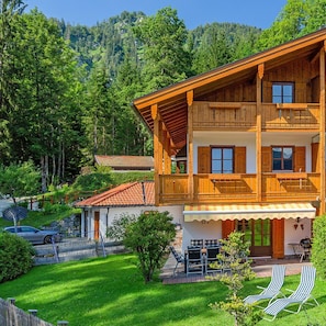 A two-story wooden chalet with a balcony, a covered patio area, and a view of a forested hillside.