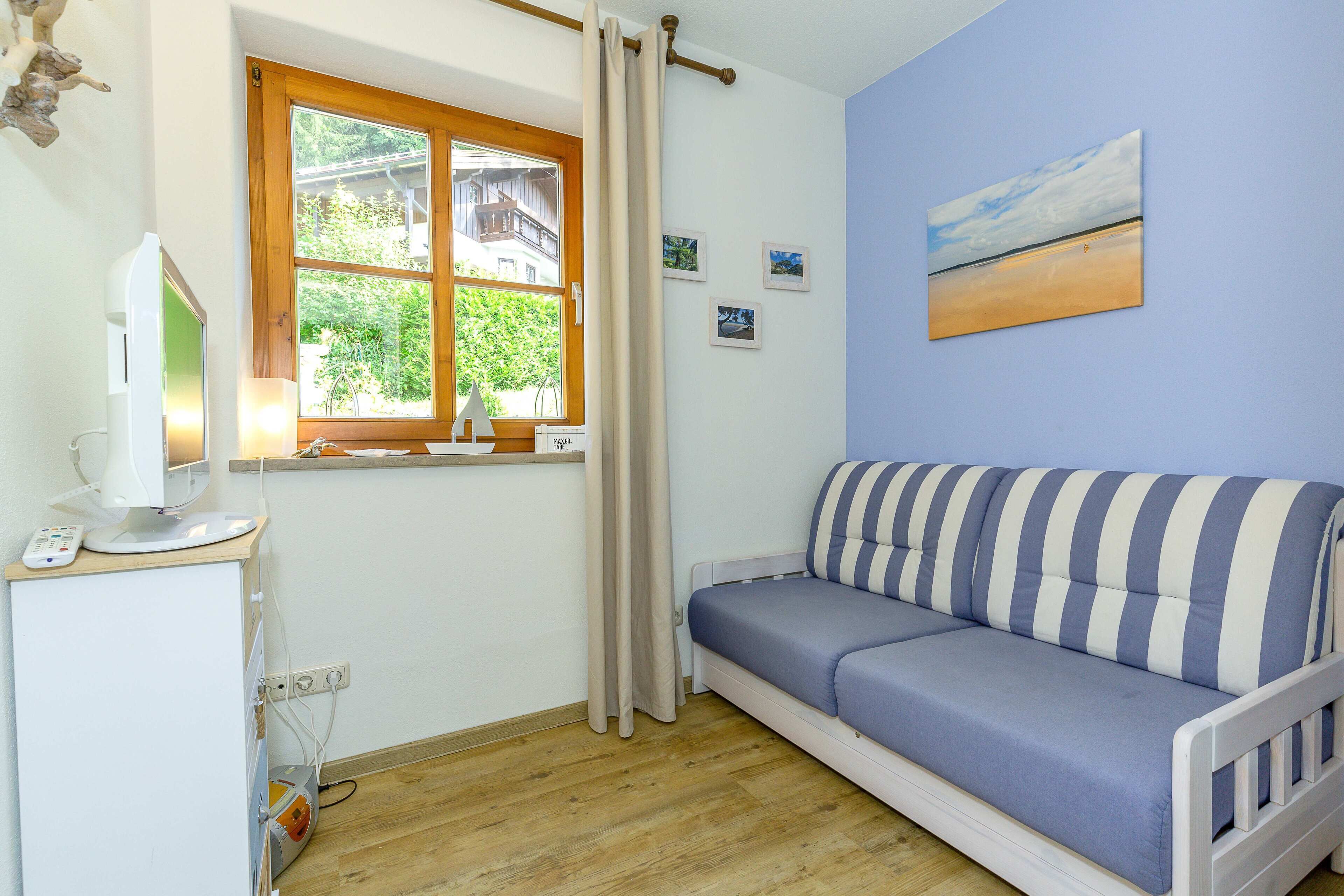 Ein Zimmer mit Holzboden, einem weißen Sofa mit gestreiften Kissen, einem kleinen Tisch mit Lampe und einem Fenster mit Vorhängen.