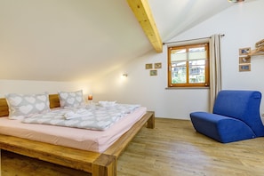 A bedroom with a wooden bed, a blue chair, and a window with a view of buildings. - Häusle Thimlefi, Lärchenweg 20 a (Reit im Winkl)