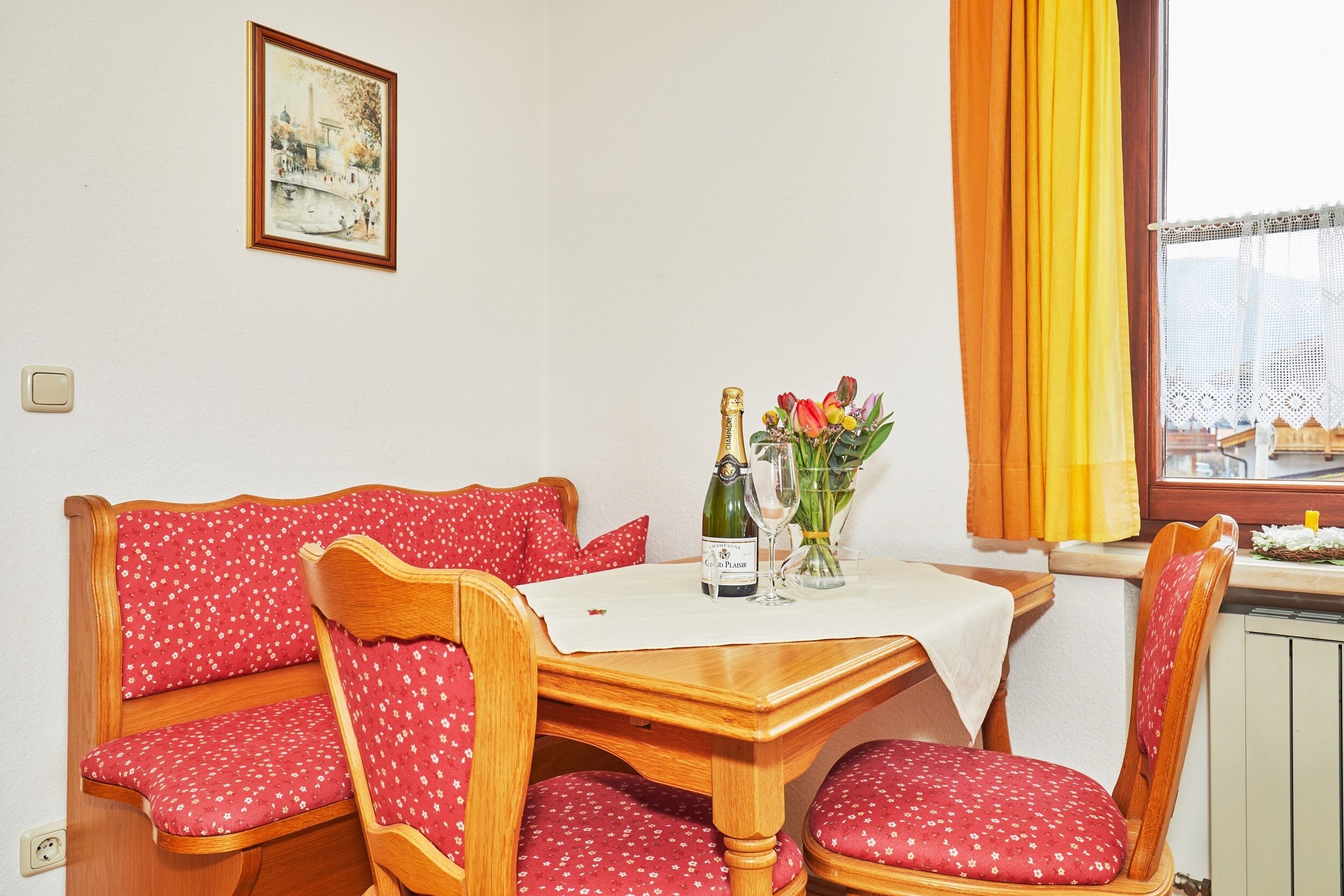 Un coin repas avec une table en bois, des chaises à motifs floraux rouges, un tableau encadré au mur et une fenêtre avec des rideaux.