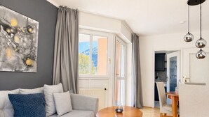 A modern living room with a grey sofa, a round wooden table, and a view of the outdoors through large windows.