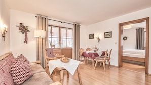 A cozy living room with a sofa, dining table, and chairs. There is a window with curtains, a lamp, and wall decorations.
