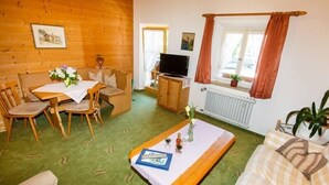 Um quarto acolhedor com piso de madeira, um tapete verde, um sofá, uma mesa de jantar com cadeiras, uma televisão e uma janela com cortinas.