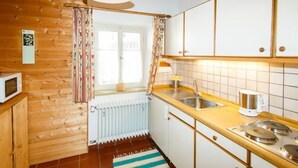 Uma cozinha compacta com paredes de madeira, uma janela com cortinas, um radiador branco, micro-ondas, pia e fogão.