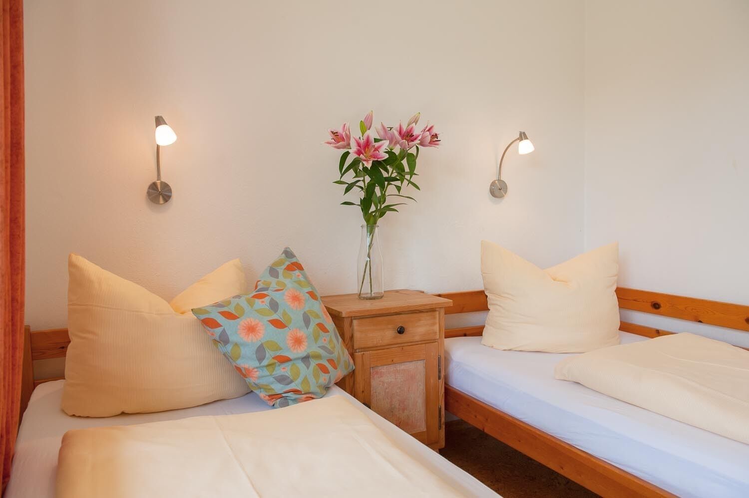 A bedroom with two beds, a nightstand, a vase with flowers, and wall-mounted lights.