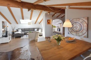 A modern living room with a wooden ceiling, a gray sofa, a dining table with a vase of flowers, and a kitchen in the background. - Apartment 3/1165, 80sqm, terrace, kitchen, 2 bedrooms and 1 living room, max 4 p. WLAN (Ruhpolding)