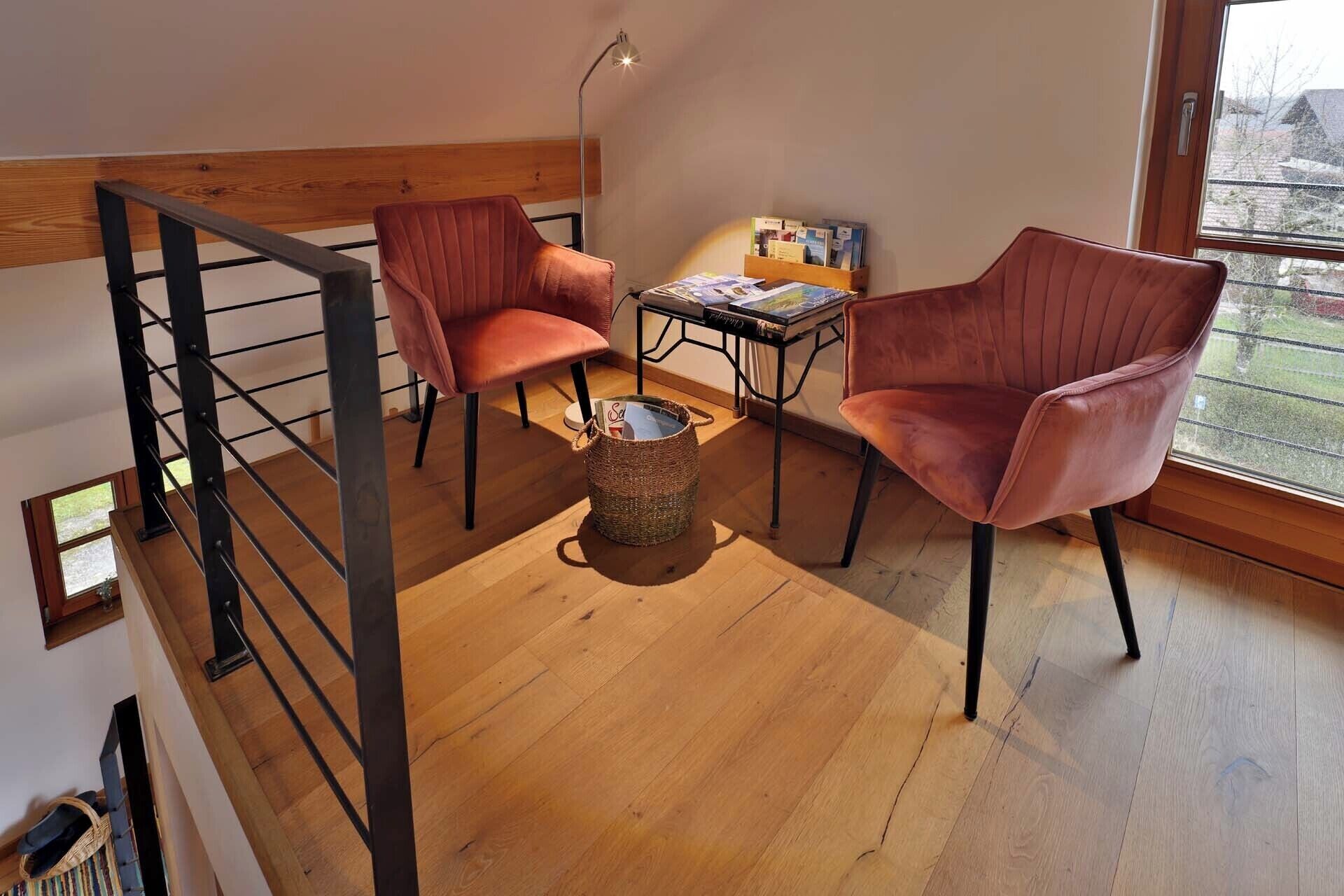 A loft-style room with a wooden floor, two red chairs, a small round table, and a bookshelf.