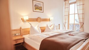 Une chambre d’hôtel avec un lit en bois, du linge de lit blanc, une table de chevet, une lampe fixée au mur et une fenêtre avec des rideaux.