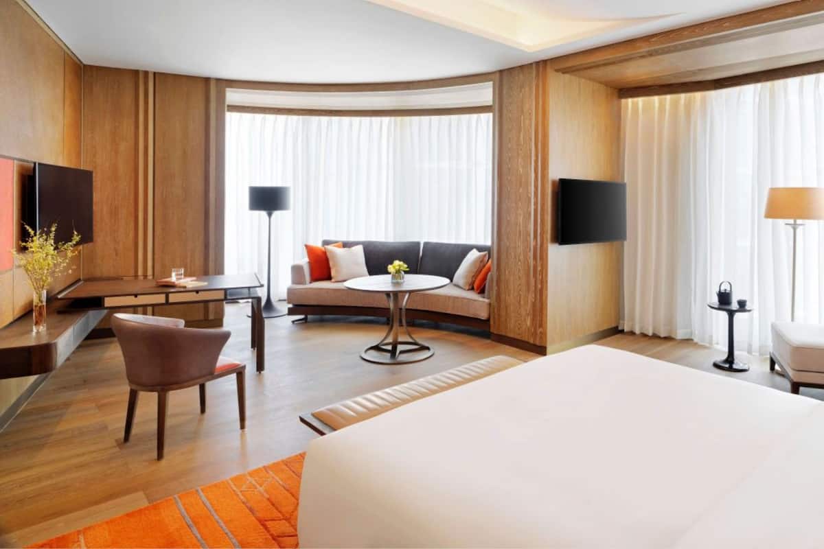 A hotel room with a large bed, a sofa, a small table, a chair, a TV, and a lamp.