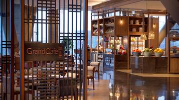 A Grand Café with a modern interior, featuring a seating area with tables and chairs, a counter with a display of fruits, and a view of the city through large windows.