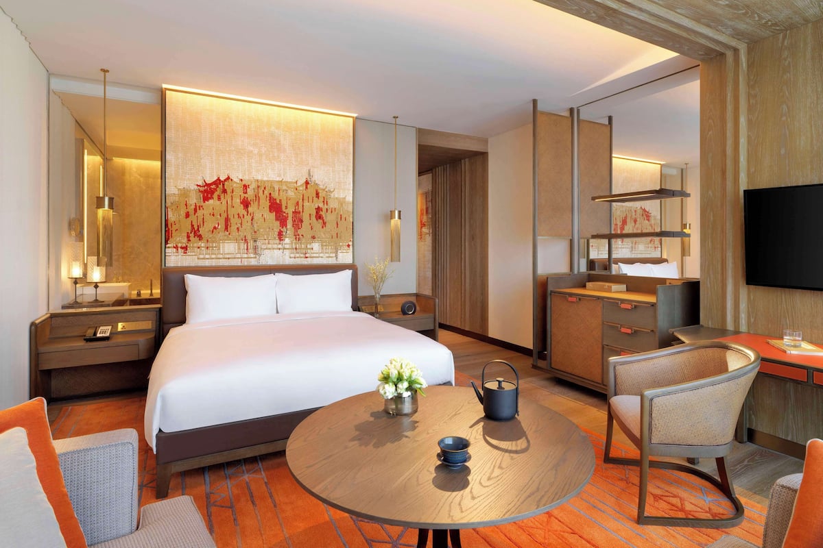 A hotel room with a large bed, a round table with a teapot and cup, a desk with a chair, a TV, and a decorative wall piece.