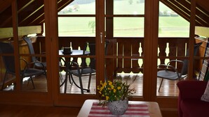 Ein Holzinnenraum mit Tisch und Stühlen, einer Vase mit gelben Blumen und Blick auf ein grünes Feld durch offene Türen.