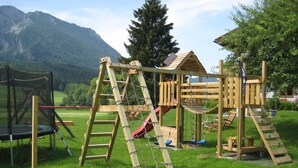 Ein hölzerner Spielplatz mit Rutschen und Klettergerüsten vor einer Berg- und Grünlandschaft.
