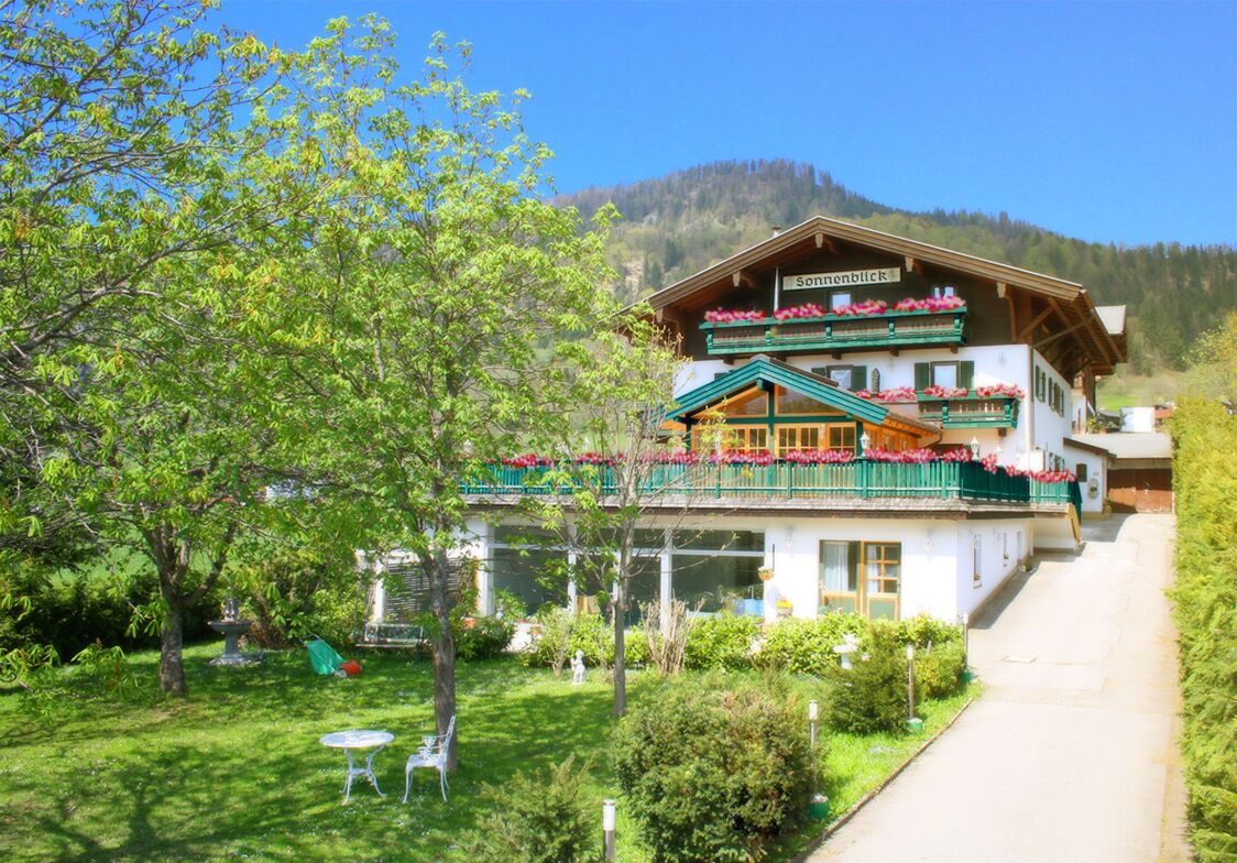 A traditional Alpine-style building with a balcony adorned with flowers, surrounded by lush greenery and a mountain backdrop.