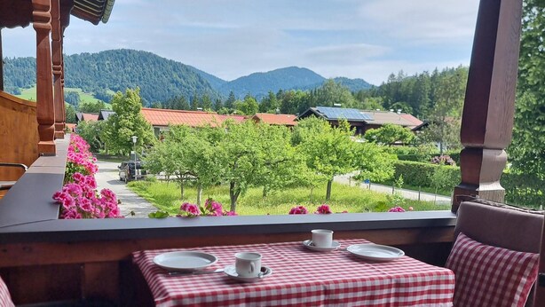 Een terras met een geruit tafelkleed, een tafel en stoelen, met uitzicht op een wijngaard en bergen.