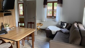 Una habitación con una mesa de comedor de madera, un sofá, un televisor montado en la pared y una ventana con cortinas.
