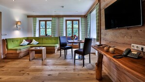 Una sala de estar moderna con un sofá verde, una mesa de centro de madera y un comedor con mesa y sillas. Un televisor está montado en la pared, y hay un escritorio de madera con un teléfono.