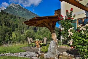 Um pátio com mesa e cadeiras de madeira, rodeado por um jardim com flores e um pequeno lago.