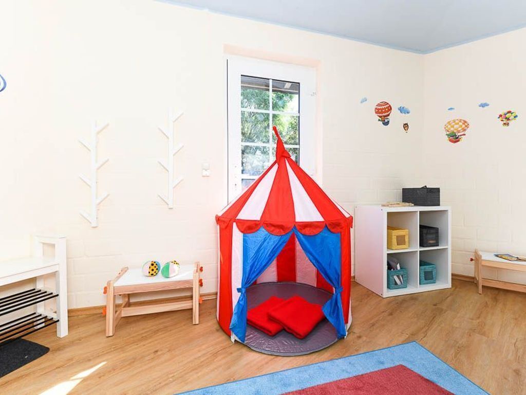Un cuarto de juegos infantil con una tienda de campaña roja y blanca, una mesita llena de juguetes y una estantería.