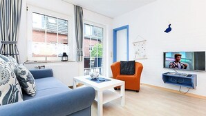 En moderne stue med en blå sofa, et hvidt sofabord og en orange lænestol. Der er et fjernsyn monteret på væggen og et vindue med gardiner.