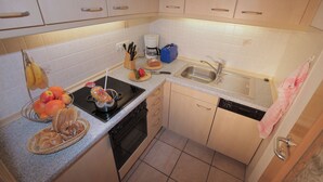 A compact kitchen with a stove, sink, and dishwasher. There are fruits and a bread basket on the counter. - Ferienwohnung 4, 2 Personen, 50 qm., 1 Schlafzimmer, DU/WC, TV, Zimmersafe, Balkon (Oberwössen)