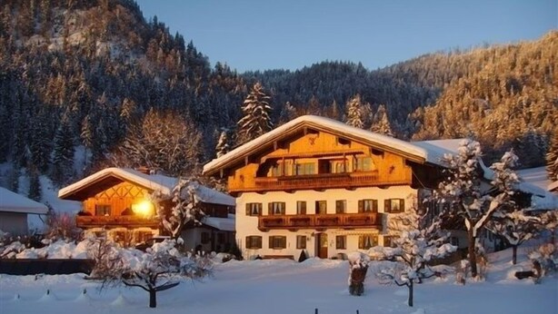A large, multi-story chalet with a wooden exterior, surrounded by snow-covered trees and a mountainous backdrop. - Morgensonne one-room app. 28sqm, shower/WC, kitchenette, balcony (Reit im Winkl)