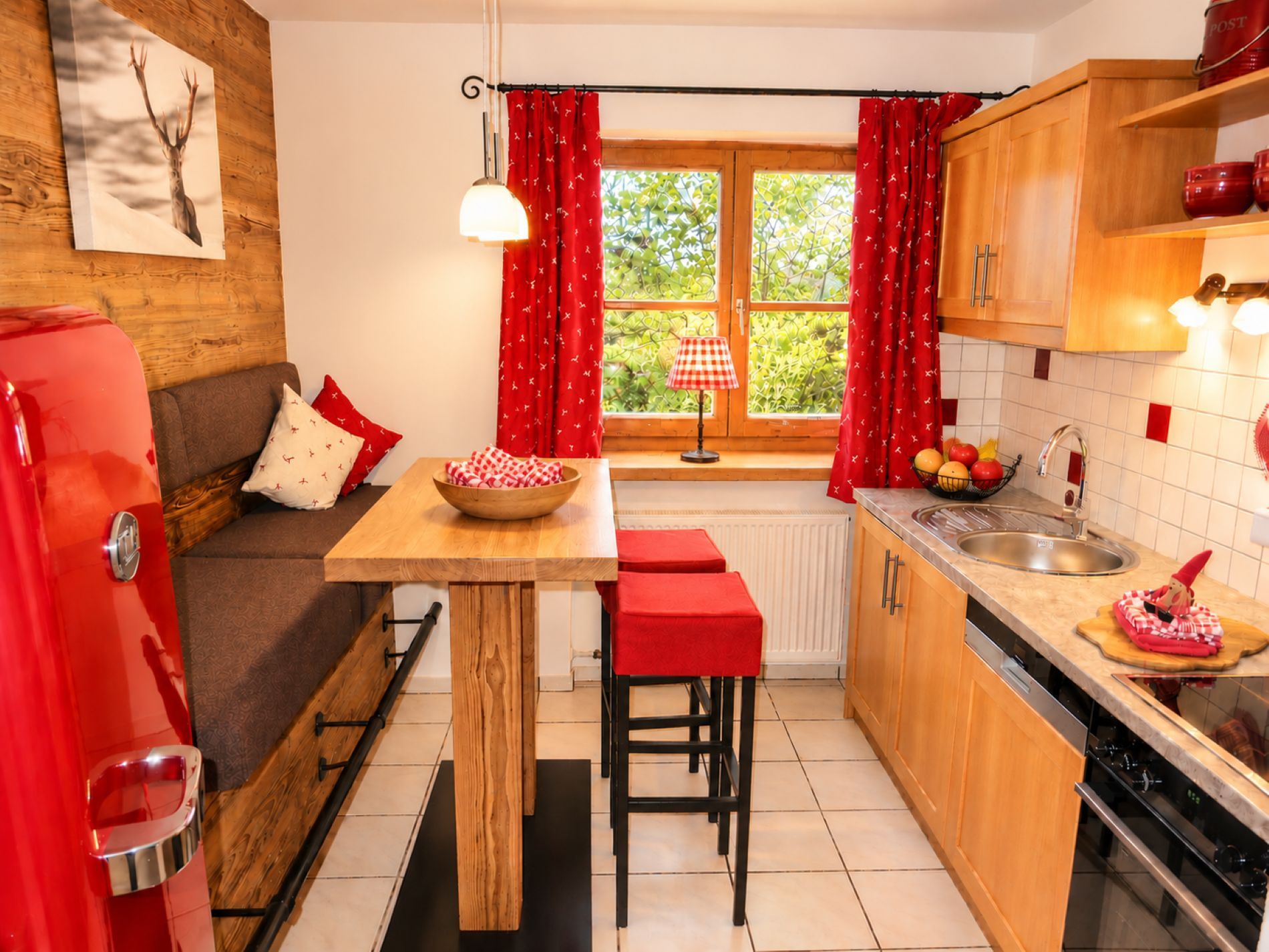 A compact kitchen with a wooden table, red accents, a refrigerator, and a window with curtains.