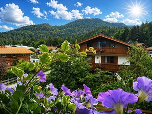 A wooden house with a balcony, surrounded by greenery and mountains.