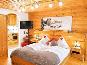 Une chambre d’hôtel avec un intérieur en bois, un lit avec une literie à motifs, un bureau avec une chaise, une télévision et un tableau représentant un paysage enneigé.