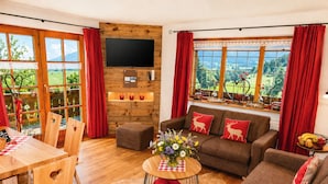 Ein Wohnzimmer mit Holzboden, einer Couch mit roten Kissen, einem Couchtisch mit Blumenvase und einem an einer Holzwand befestigten Fernseher.